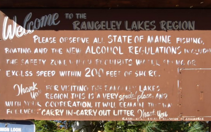 A sign reads, "Welcome to the Rangeley Lakes Region," along with other text, much of which is unreadable due to wear. 